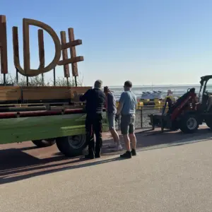Arbeiter verladen große Holzbuchstaben an einer Strandpromenade am Meer.