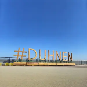 Großer hölzerner Schriftzug #DUHNEN vor klarem blauem Himmel.