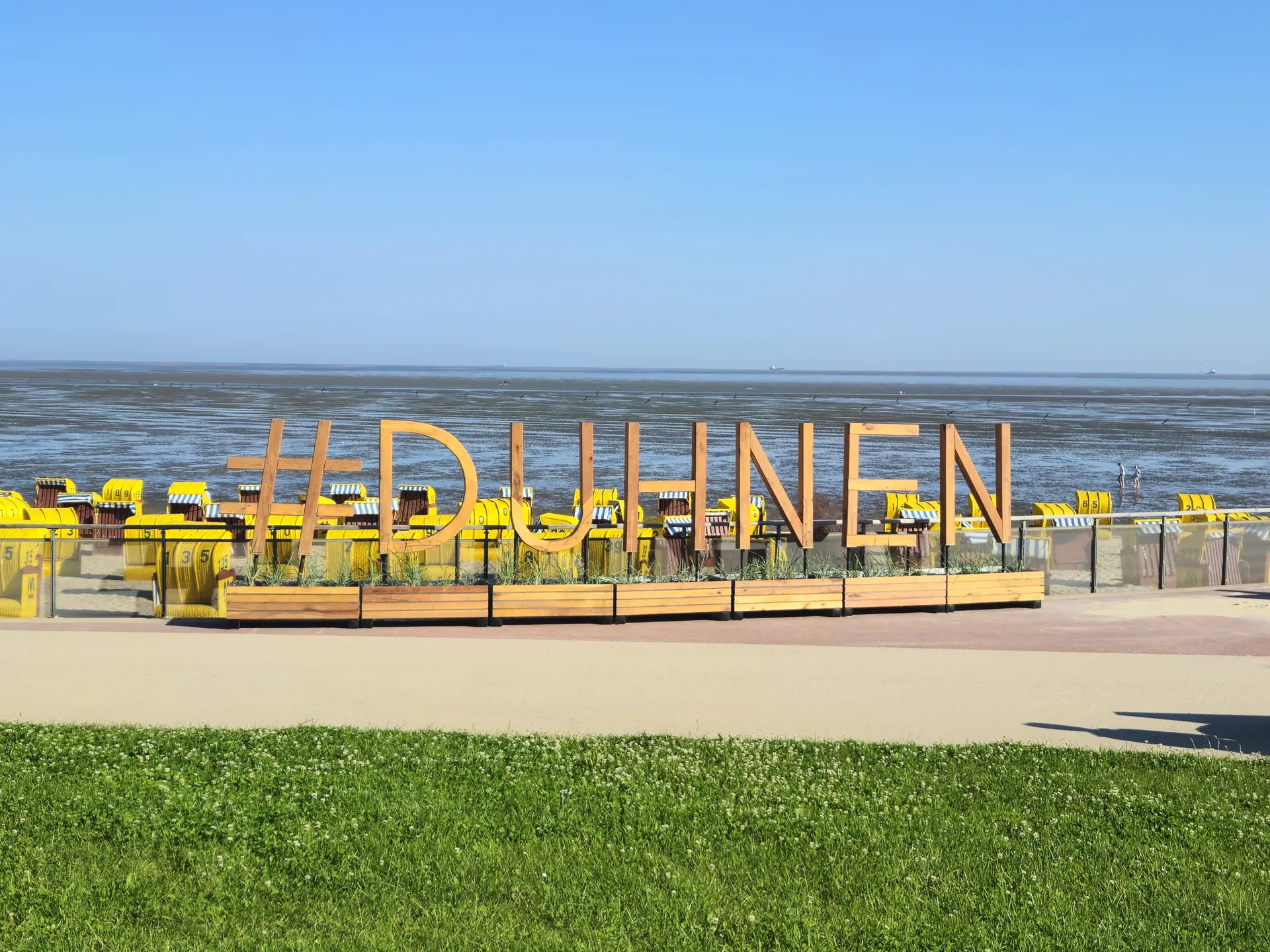 Holzschriftzug #DUHNEN am Strand mit gelben Strandkörben vor Wattenmeer.